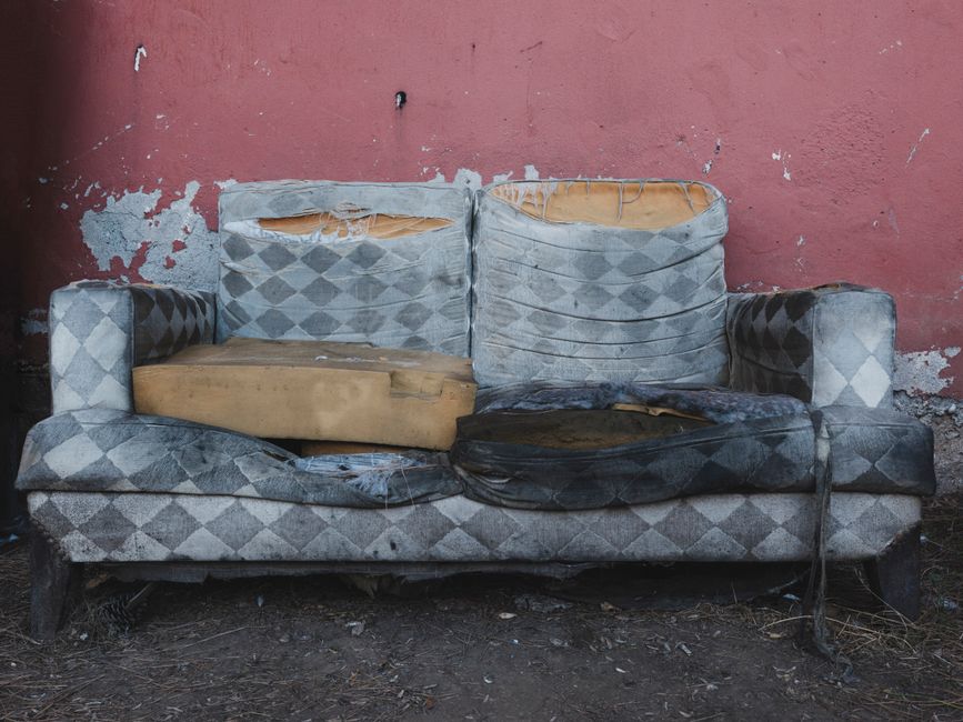 A worn-out, damaged fabric sofa with a diamond-patterned upholstery in shades of grey, situated outdoors against a weathered, peeling pink wall. The sofa is visibly stained and torn, with sections of 