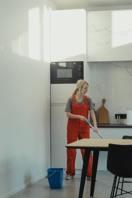 A woman wearing orange overalls and a grey t-shirt is performing surface cleaning using a mop in a modern kitchen. The kitchen features white marble walls with subtle grey veining, a built-in microwav