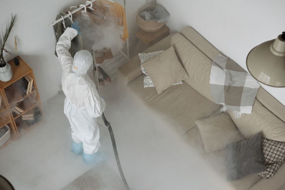 A cleaning professional from 0800carpetcleaner.co.uk is performing surface cleaning and sanitisation in a living room. They are wearing a white protective suit, mask, gloves, and shoe covers, and usin