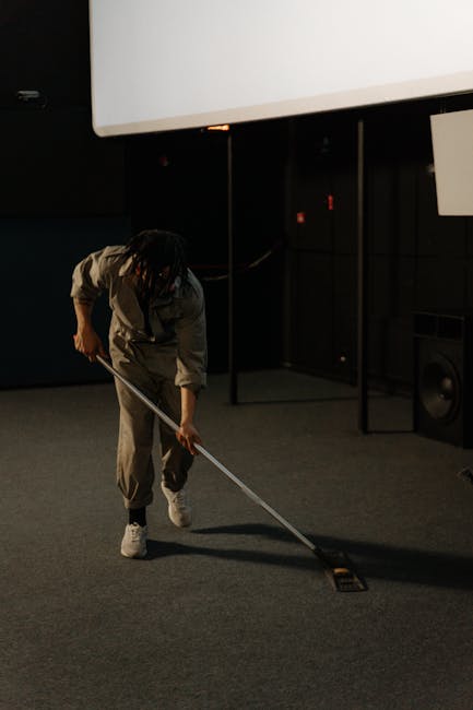 A professional cleaner dressed in white protective clothing and a face mask is operating a fogging or steam cleaning machine over a beige carpet in a living room. The room features a cream sofa with b