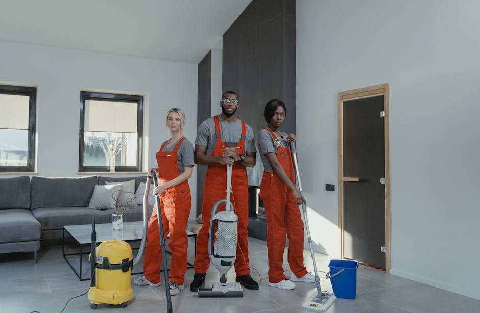 A professional cleaner from 0800carpetcleaner.co.uk is standing on a modern, light-colored tiled floor in a well-lit room with large glass sliding doors, which reveal an outdoor balcony. She is wearin
